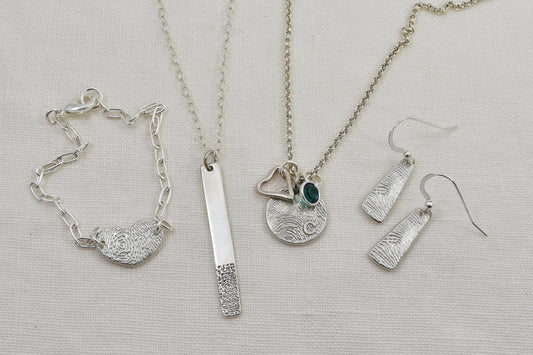 Fingerprint Jewelry Flat Lay Includes a Heart Fingerprint Bracelet, a fingerprint bar necklace, a circle fingerprint necklace accessorized with an open heart symbol charm and a birthstone charm and a pair of fingerprint paddle earrings.