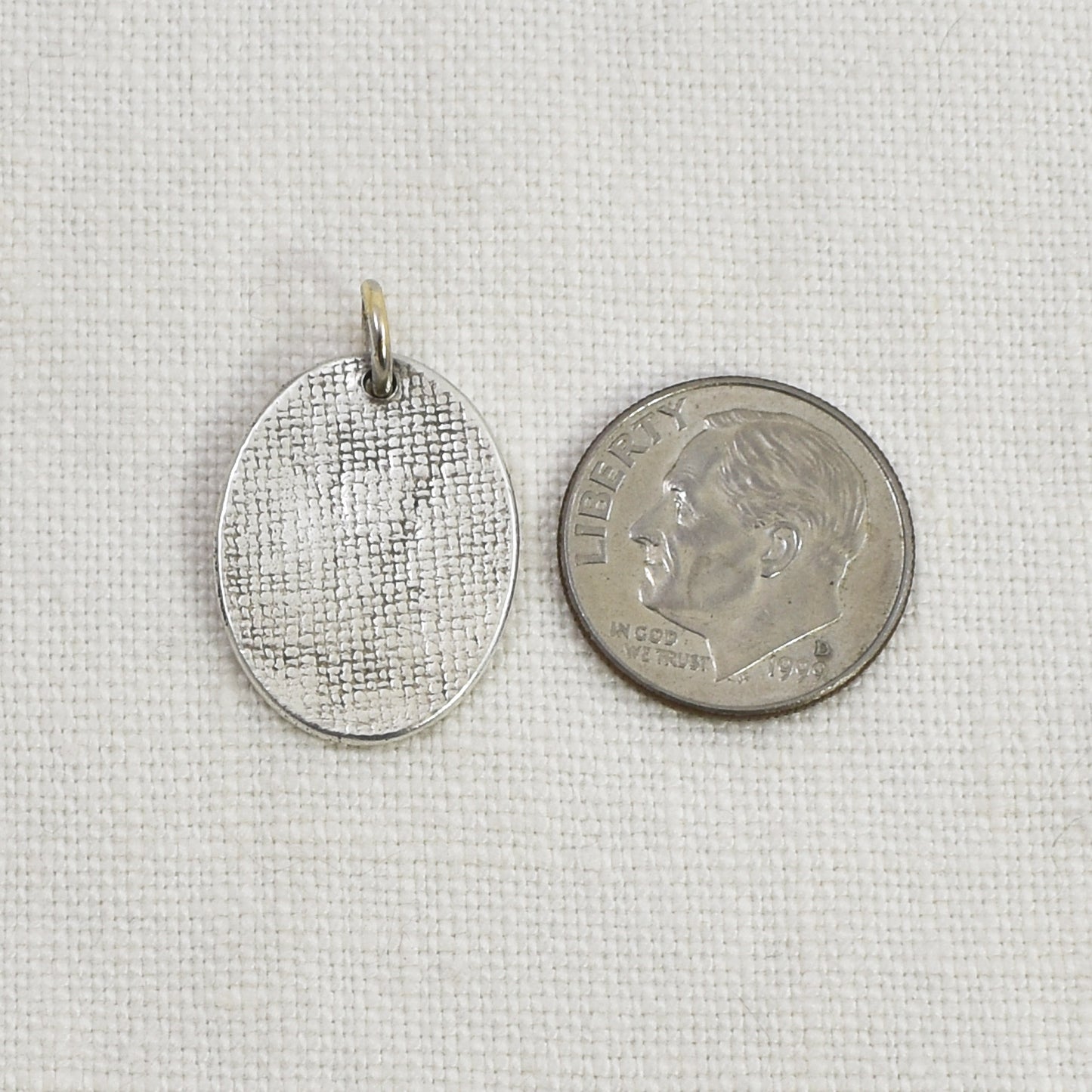 Back of a Silver oval pendant next to a U.S. dime on a light colored linen background