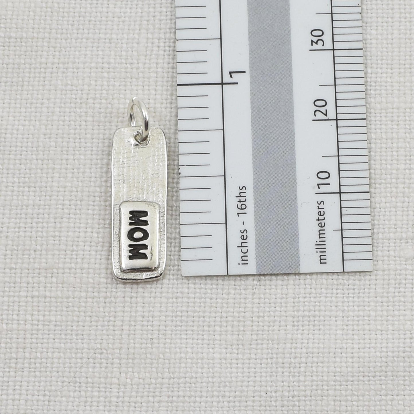 Back of silver fingerprint pendant showing 'MOM' engraving next to a ruler for scale