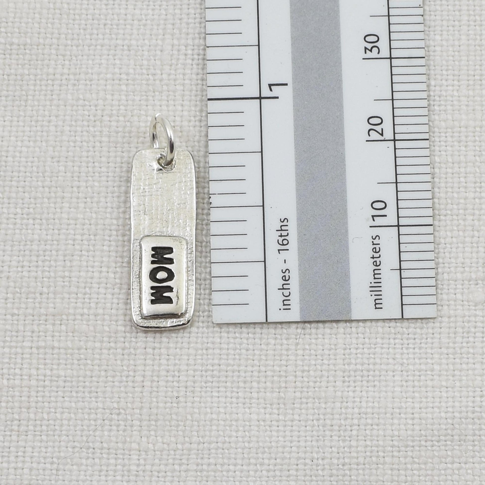 Back of silver fingerprint pendant showing 'MOM' engraving next to a ruler for scale