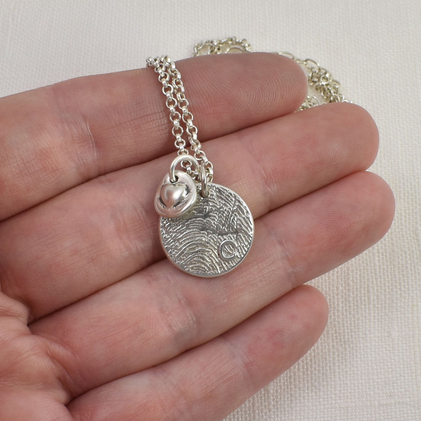 Tiny Sterling Silver Heart Charm shown next to a five-eighths inch Circle Fingerprint Pendant hanging on a sterling silver rolo chain