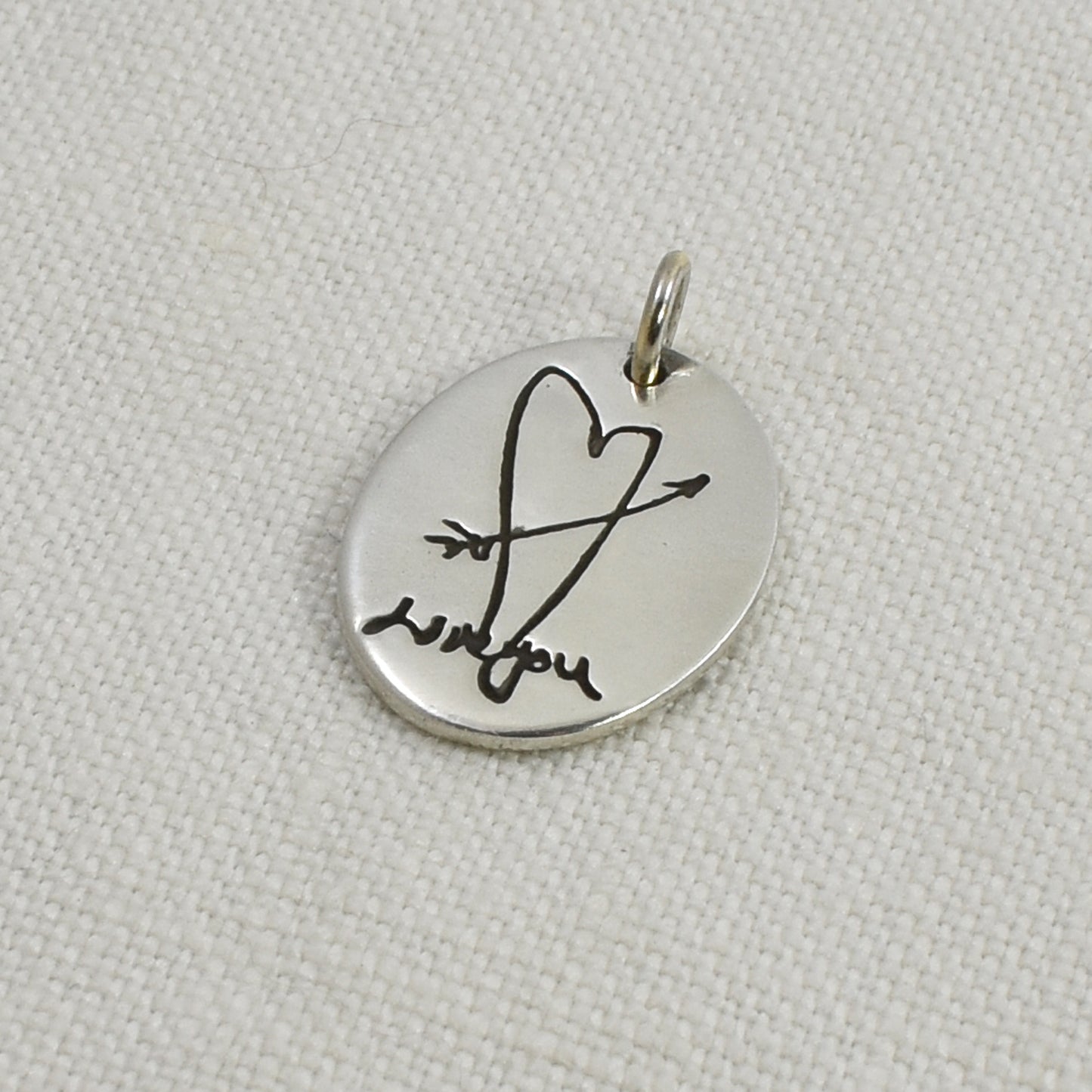 Sterling Silver Oval Pendant engraved with a hand drawn heart and handwriting photographed on a linen background.  