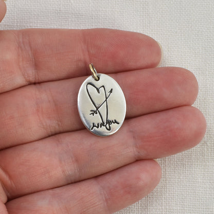 Silver pendant with heart and arrow design held between fingers on a light fabric background